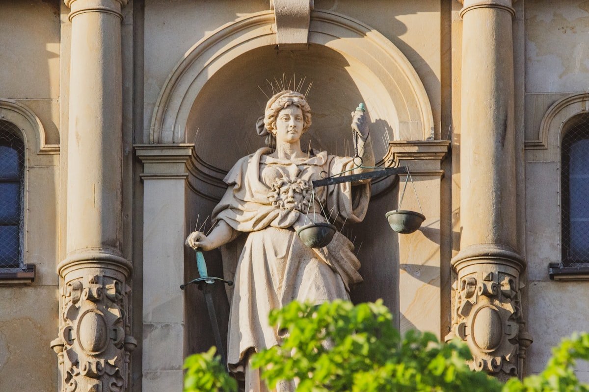 A seated stone figure of Justice holding scales, surrounded by foliage.